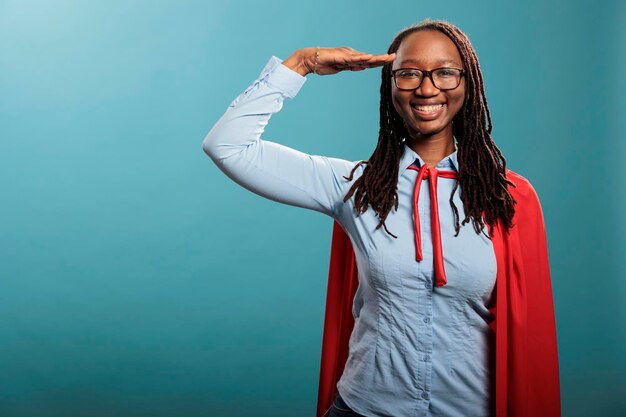 Patriotic justice defender saluting with respect while standing on blue background while smiling at camera. Brave and proud happy superhero woman wearing mighty hero cloak while making soldier salute.