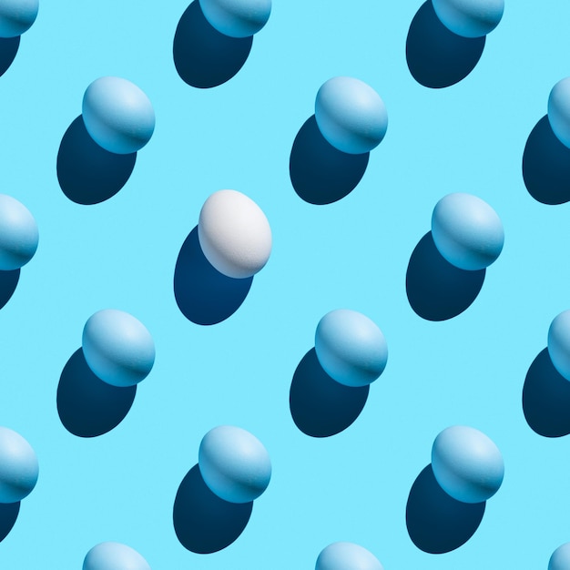A pattern of blue eggs and one white with a hard shadow on a blue background. The concept of minimalistic Easter backgrounds.