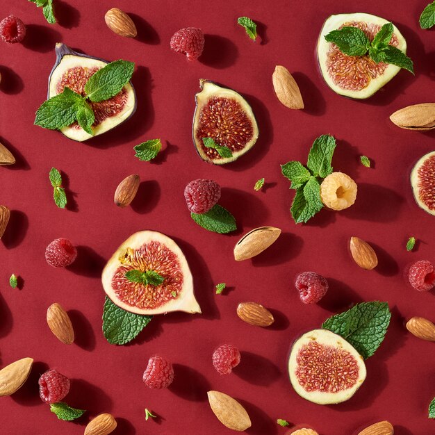 濃い赤の背景にイチジク、ミントの葉、ラズベリー、アーモンドの健康的な半分のパターン。食品の背景。フラットレイ