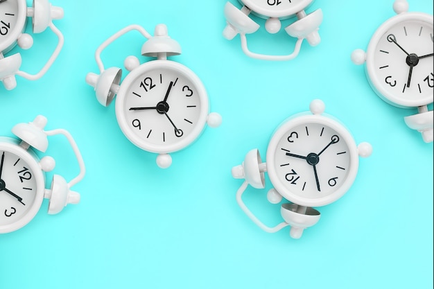 A pattern of many white classic alarm clocks in the form of a pattern on a blue background. Top view with a copy of the space, flat lay.