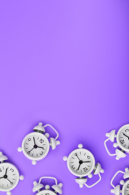 A pattern of many white classic alarm clocks in the form of a pattern on a purple background. Top view with a copy of the space, flat lay.