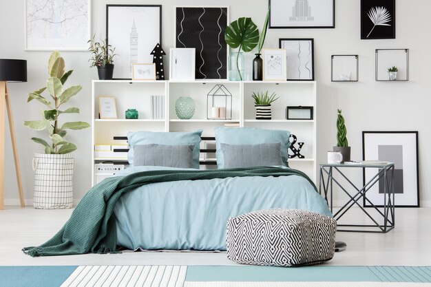 Photo patterned pouf in front of blue bed with green blanket in bedroom interior with ficus and posters