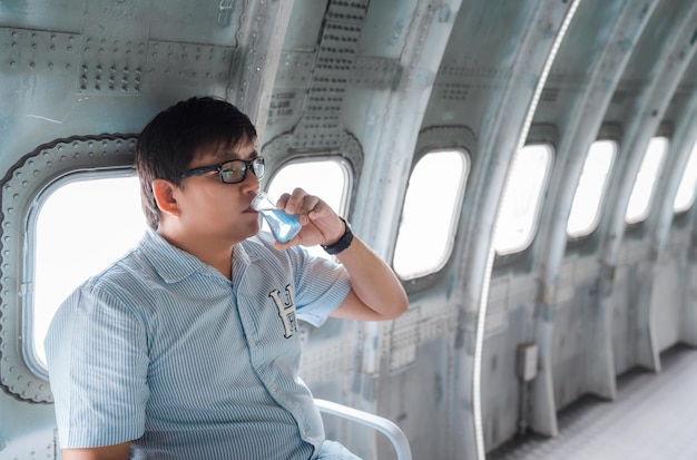 L'uomo asiatico obeso paunchy beve liquido blu dalla bottiglia di matraccio erlenmeyer su un aeroplano d'acciaio vicino alla finestra