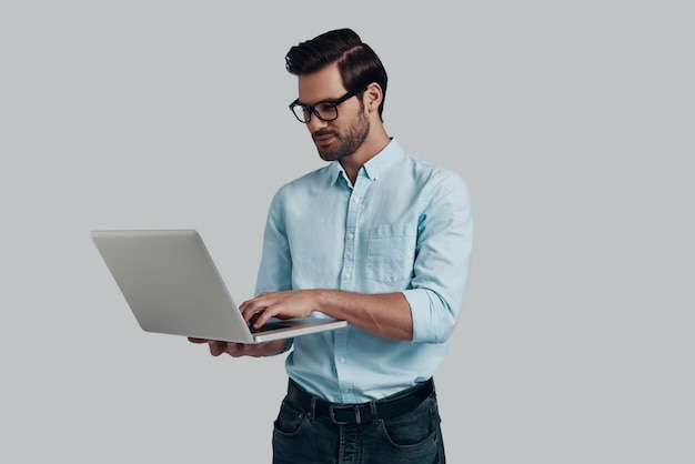 Paying attention to every detail. Young man using laptop while standing against grey background