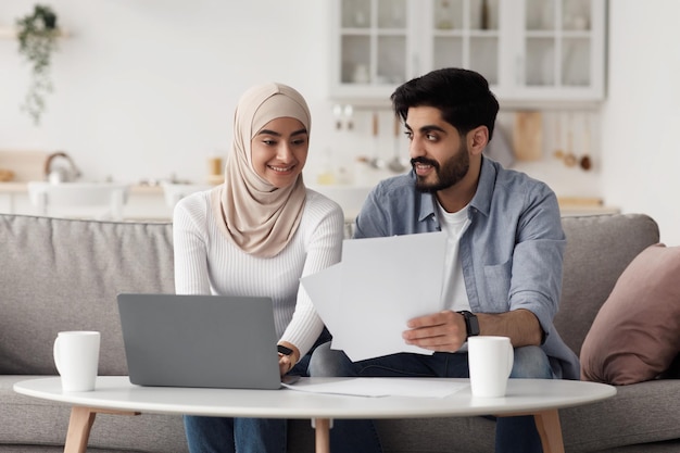 Payment of bills, taxes, finance, family budget and bookkeeping at home, covid-19. Smiling millennial arab lady in hijab and man working with documents and laptop in living room interior, copy space