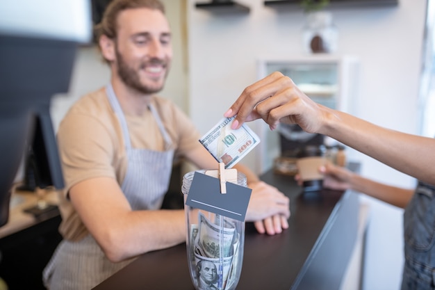 Payment for order. Womans hand holding out hundred dollar bill for coffee and smiling man in apron behind counter