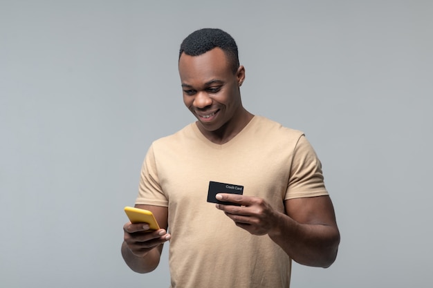 Payment transaction. Smiling young adult african american man with smartphone and bank card in hands standing against light background