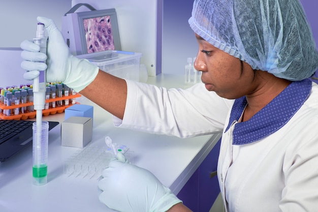 PCR test for coronavirus. African young female tech, scientist or medic in white coat and hat performs test to detect SARS-CoV-2 in patient samples, mouth swabs. Automatic pipette in gloved hands.