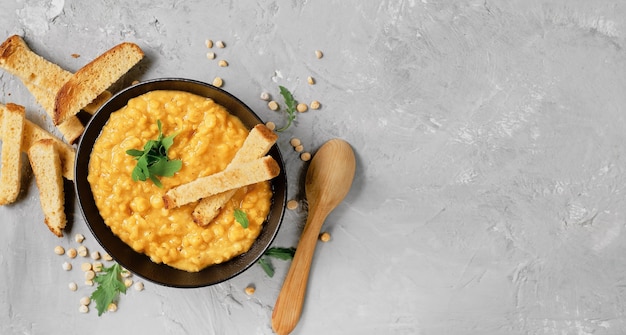 Pea soup with vegetables and spices in a clay bowl, next to a wooden spoon. Top view of hot yellow pea soup, focusing on the top of the dish. Layout on concrete gray table with copy space