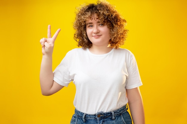 Peace gesture Body positive Healthy nutrition Self esteem Cheerful lovely smiling overweight woman with curly hair showing hippie friendly hand symbol isolated on yellow