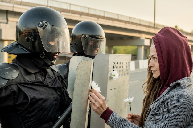 Peace-loving young woman in hood standing in front of police force attaching daisies to riot shields at rebellion