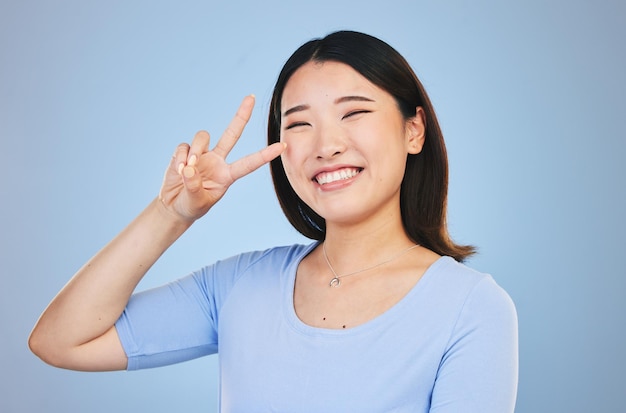 Peace sign and portrait of woman with hand for emoji in studio blue background with gen z style fashion or happiness Face smile and Asian model with gesture expression