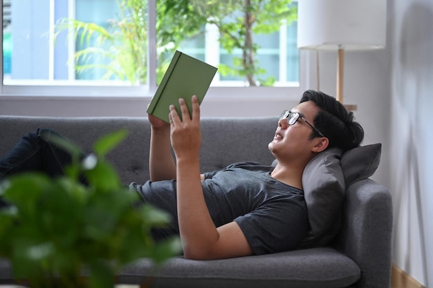 Peaceful Asian man lying on couch and reading book