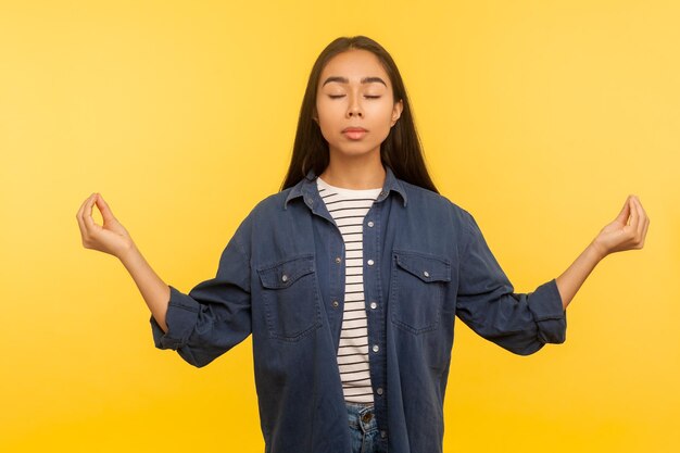 Peaceful mind. Portrait of woman in denim shirt meditating with mudra gesture and feeling harmony balance, taking break to relax, concentrate thoughts. indoor studio shot isolated on yellow background