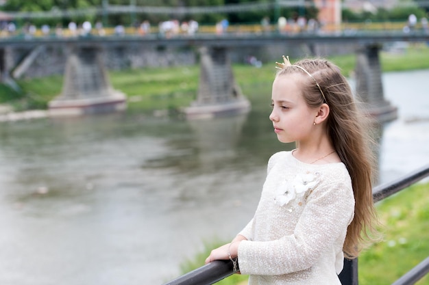 Peaceful princess. Girl princess with little crown river background copy space. Princess girl with long hair in white clothing in pensive mood. Happy childhood. Thoughtful lonely child at riverside.