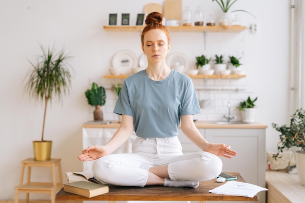 Peaceful redhead young woman is sitting with closed eyes on table in lotus position and makes deep breathexhalation at home office Peaceful female sitting in lotus pose