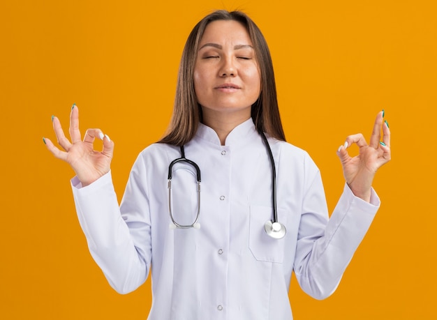 Peaceful young asian female doctor wearing medical robe and stethoscope meditating with closed eyes isolated on orange wall