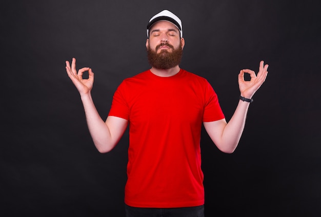 peaceful young bearded man in red t-shirt making zen gesture over black