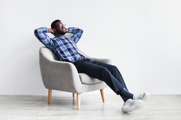 Peaceful young black man relaxing in armchair against white studio wall free space