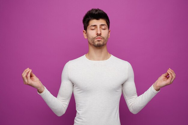 Peaceful young handsome man doing yoga gesture with closed eyes on purple background