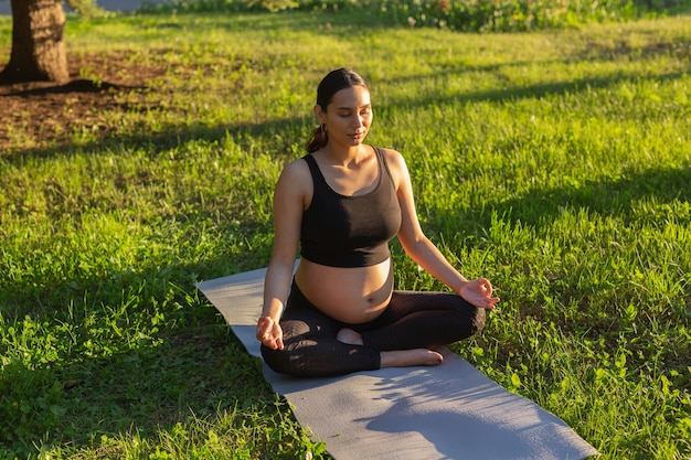 Peaceful young positive pregnant woman in gymnastic suit does yoga and meditate sitting on mat on