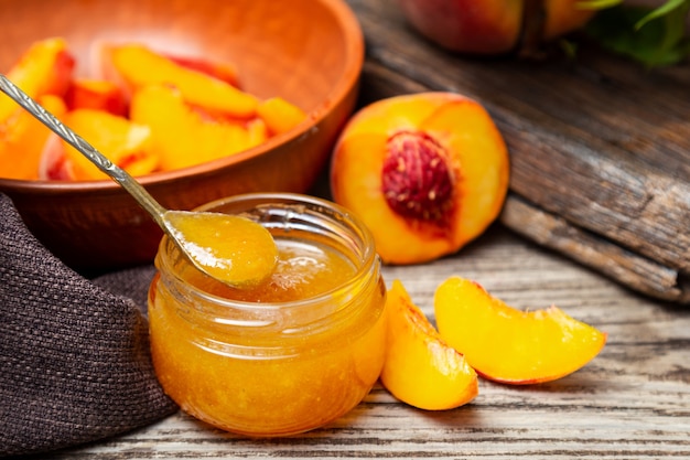 Peach jam in glass jar with peach wedges and whole peach fruit. Peach jam on wooden table.