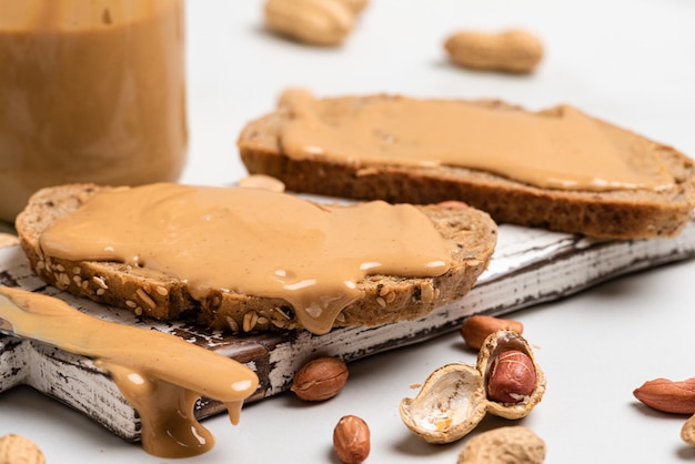 Peanut butter in a glass jar, peanuts, kitchen knife and peanut butter sandwiches on white surface