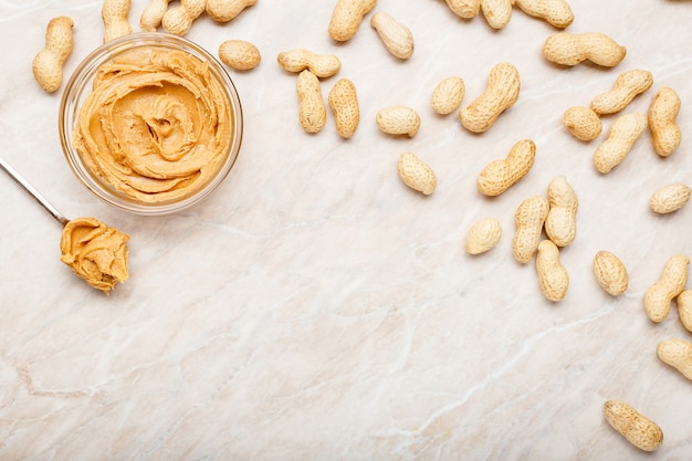 Peanut butter in glass plate with peanuts in shell, spoon with peanut butter. Creamy peanut paste flat lay with place for text on white marble background for cooking breakfast.