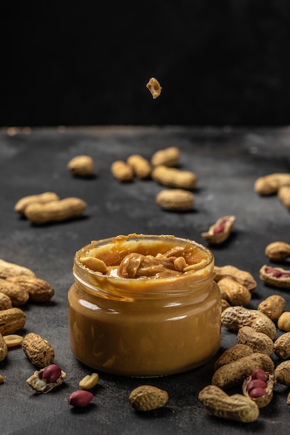 Peanut butter or paste in an open jar and peanuts in the peel scattered on dark background American dessert concept vertical image top view place for text