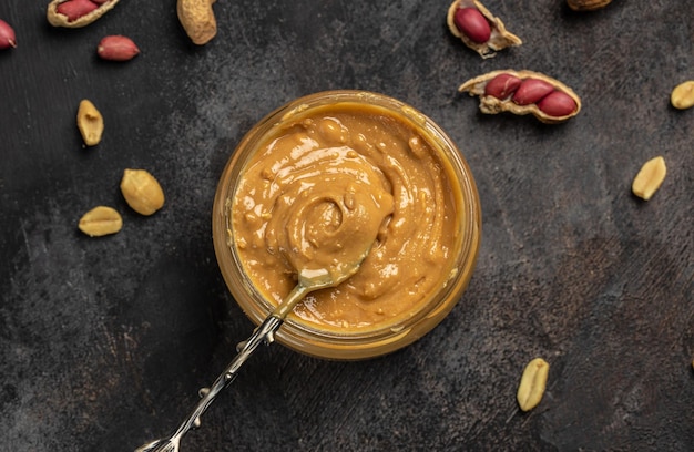 Peanut butter or paste in an open jar and peanuts in the peel scattered on a dark background banner menu recipe place for text top view