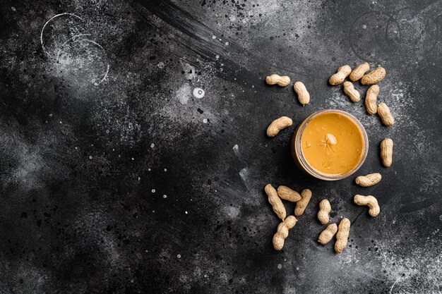 Peanut paste in an open jar and peanuts set on black dark stone table background top view flat lay with copy space for text