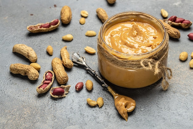 Peanut paste in an open jar in spoon near creamy peanut butter peanuts scattered on a light background place for text top view