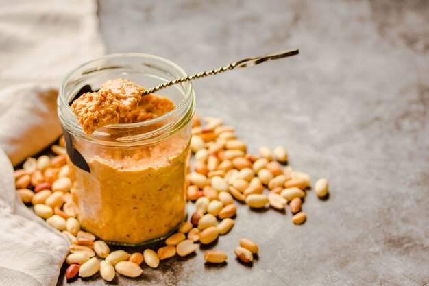 Peanut paste in an open jar and and spoon, peanuts on the gray table.