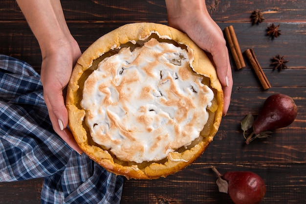 Pear pie with cream in female hands, homemade pastries on wooden table, with spices. Copy space.