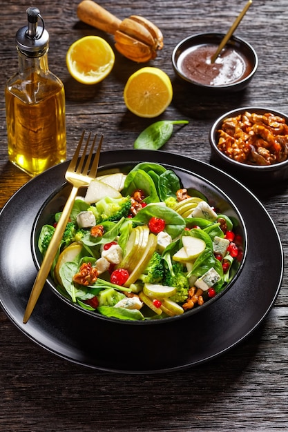 Pear salad with blue cheese cubes, broccoli, spinach leaves, caramelized walnuts and pomegranate seeds  on a black plate on wooden table, thanksgiving side dish, vertical view from above