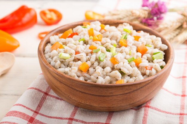 Pearl barley porridge with vegetables in wooden bowl on a white wooden surface and linen textile. Side view, selective focus.