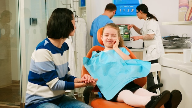 Pediatric dentist checking teeth x-ray of kid patient talking with man assistant in background. sad child showing affected mass to mother sitting stomatological chair dental unit.