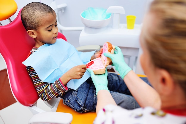 소아 치과 의사는 치아를 제대로 닦기 위해 치과 의자에 앉아있는 아프리카 계 미국인 어린이를 가르치고 있습니다. 소아 치과