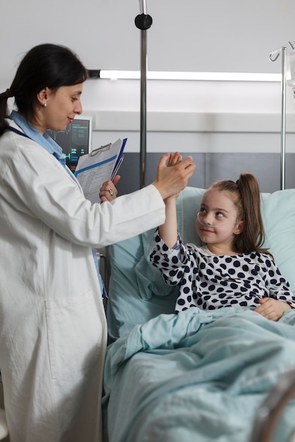 Pediatric expert doing high five gesture with ill kid sitting in patient bed. Pediatrician high fiving sick little girl under treatment resting in children healthcare facility.