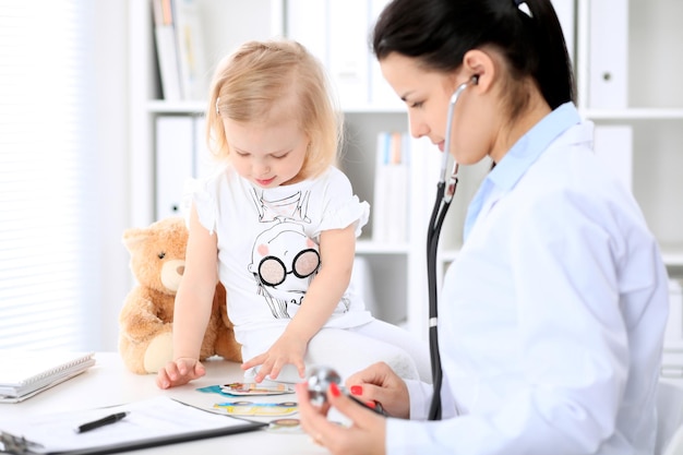 Pediatrician is taking care of baby in hospital. little girl is\
being examine by doctor with stethoscope. health care, insurance\
and help concept.