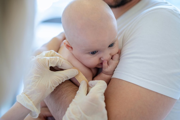 Педиатр в стерильных перчатках закрывает место укола руки ребенка лейкопластырем
