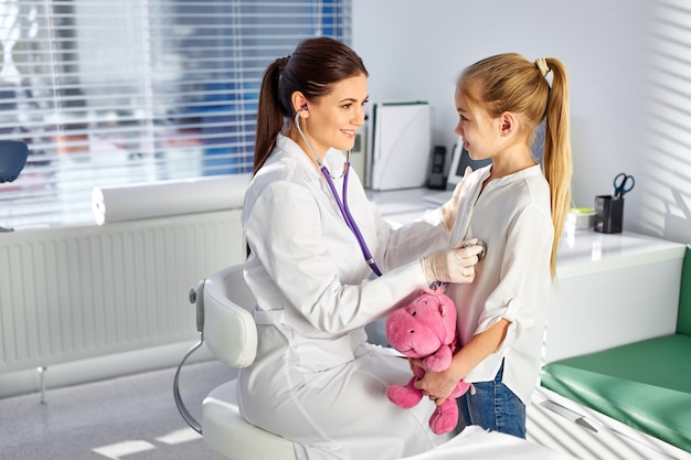 Pediatrician using stethoscope for listening lungs and heart of child girl, female doctor examines teenage girl in clinic. concept of pediatric medicine