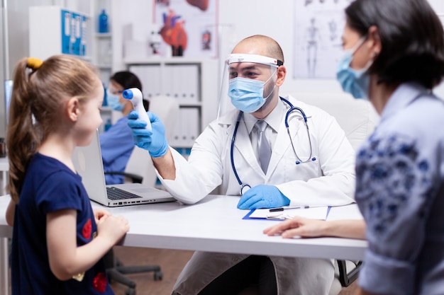 病院のオフィスで子供の体温を測定するフェイスマスクアガシントコロナウイルスを身に着けている小児科医。ヘルスケアサービスの相談を提供するヘルスドクタースペシャリスト。
