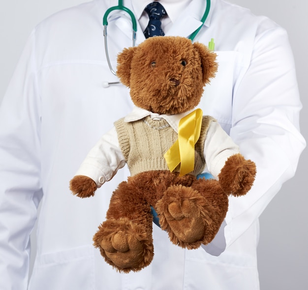 Pediatrician in white coat, blue latex gloves holds a brown teddy bear with a yellow ribbon on a sweater, concept of the fight against childhood cancer