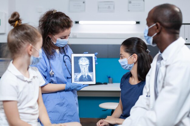 Pediatrician woman nurse holding tablet computer showing patient radiography during clinical inspection in hospital office. Medical team with protective face mask against coronavirus