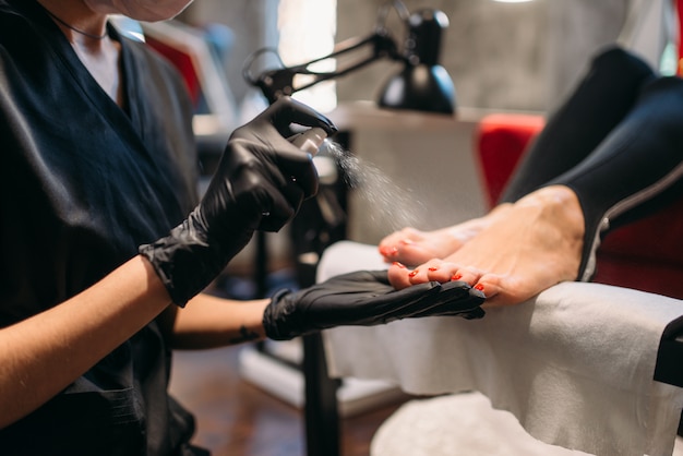 Pedicure master in black gloves sprays foot nails of female client, beauty salon. Professional  fingernail care