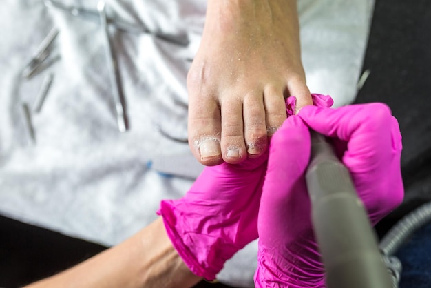 Pedicuremeester in roze handschoenen snijdt de nagels van de nagelriem en schellak in de pedicuresalon met behulp van boor Professionele pedicure in cosmetologiekliniek Hygiëne voor voeten in schoonheidssalon