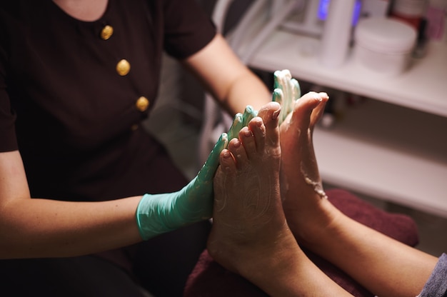 Pedicurist applying nourishing moisturizer cream on woman's legs and massaging them in pedicure room