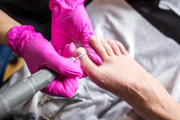 Pedicurist master in pink gloves cuts the cuticle and shellac toe nails in the pedicure salon using drill Professional pedicure in cosmetology clinic Hygiene for feet in beauty salon