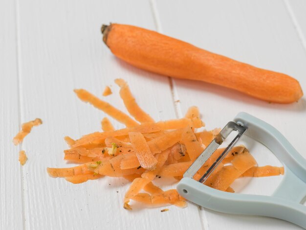 Фото Очищенная морковь и чистящие средства на белом деревянном столе. чистка моркови специальным ножом.